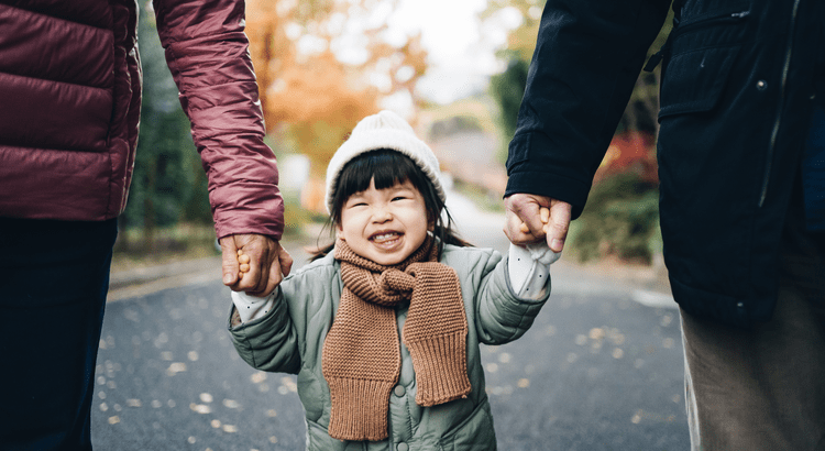 Young child smiling while holding parents’ hands during winter—symbolizing the joy and opportunity families can find when selling a home in the winter housing market.